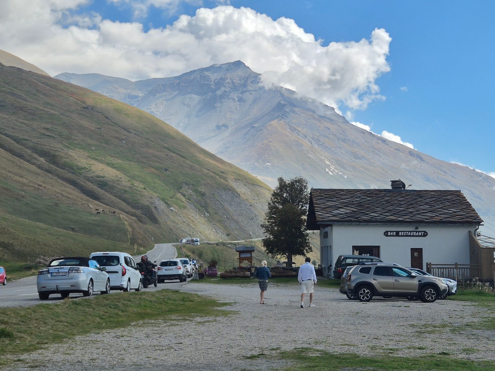 A bar restaurant with a gravel car park full of cars below a tall mountain in the Alps.