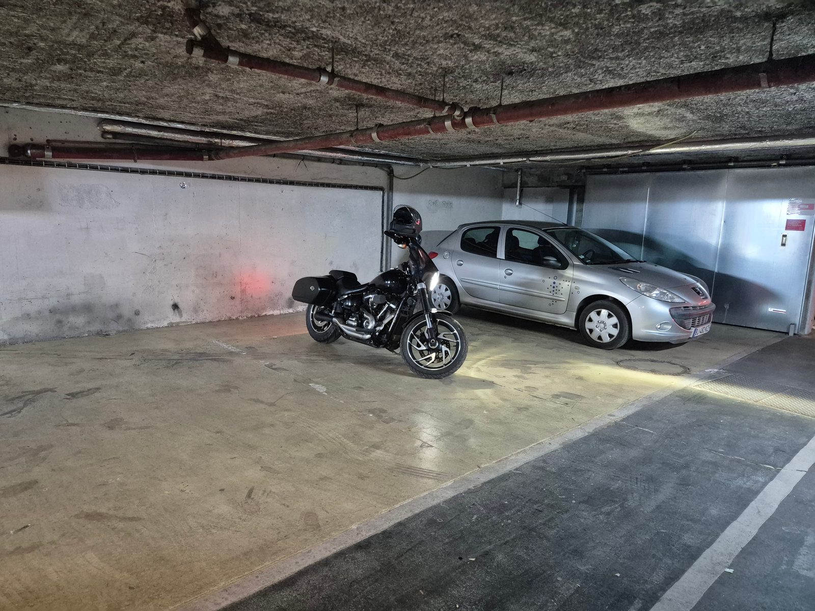 A black cruiser motorcycle with a helmet on the seat parked in an underground car park next to a silver Peugeot.