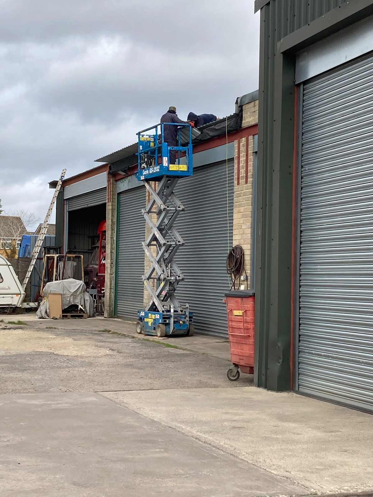 Two men on a blue scissor lift working on the roof edge of an industrial garage unit with grey roller shutter doors.
