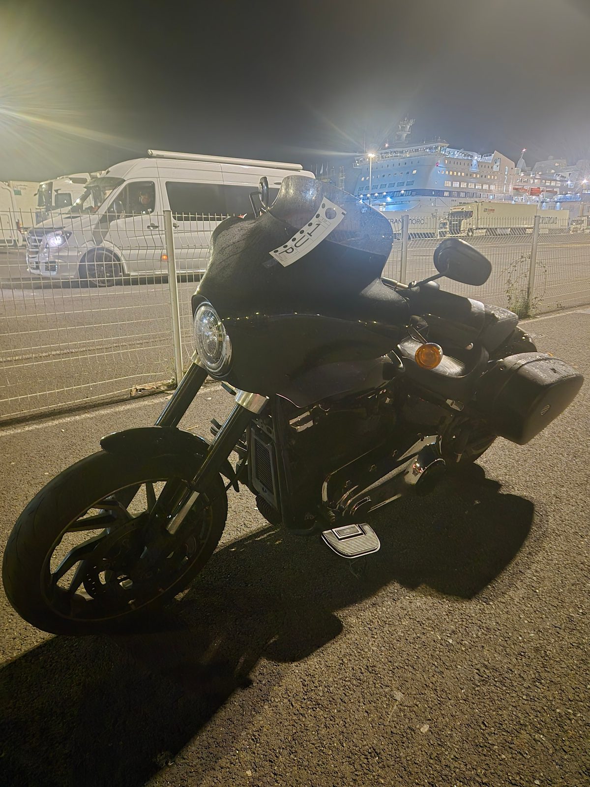 A black motorcycle parked by a fence at a ferry port at night with a large ferry lit up in the background.