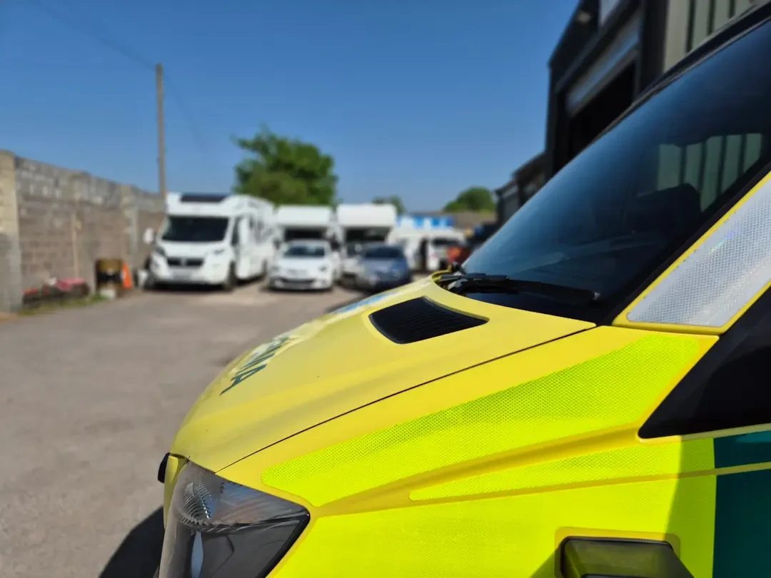 The bonnet of a yellow and green ambulance in the foreground, with white motorhomes and cars parked behind under a clear blue sky.
