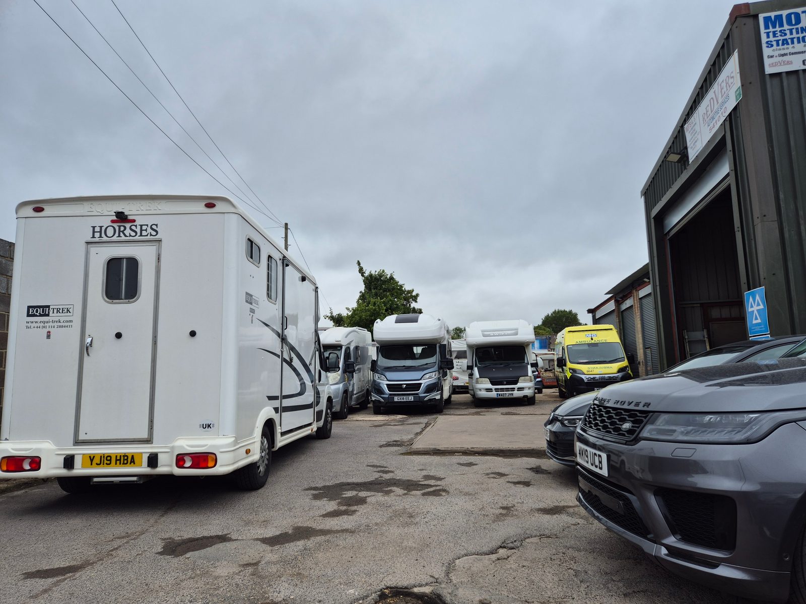 An Equi-Trek horsebox, several coachbuilt motorhomes, a yellow NHS ambulance and a grey Range Rover parked outside the Redvers MOT testing station.