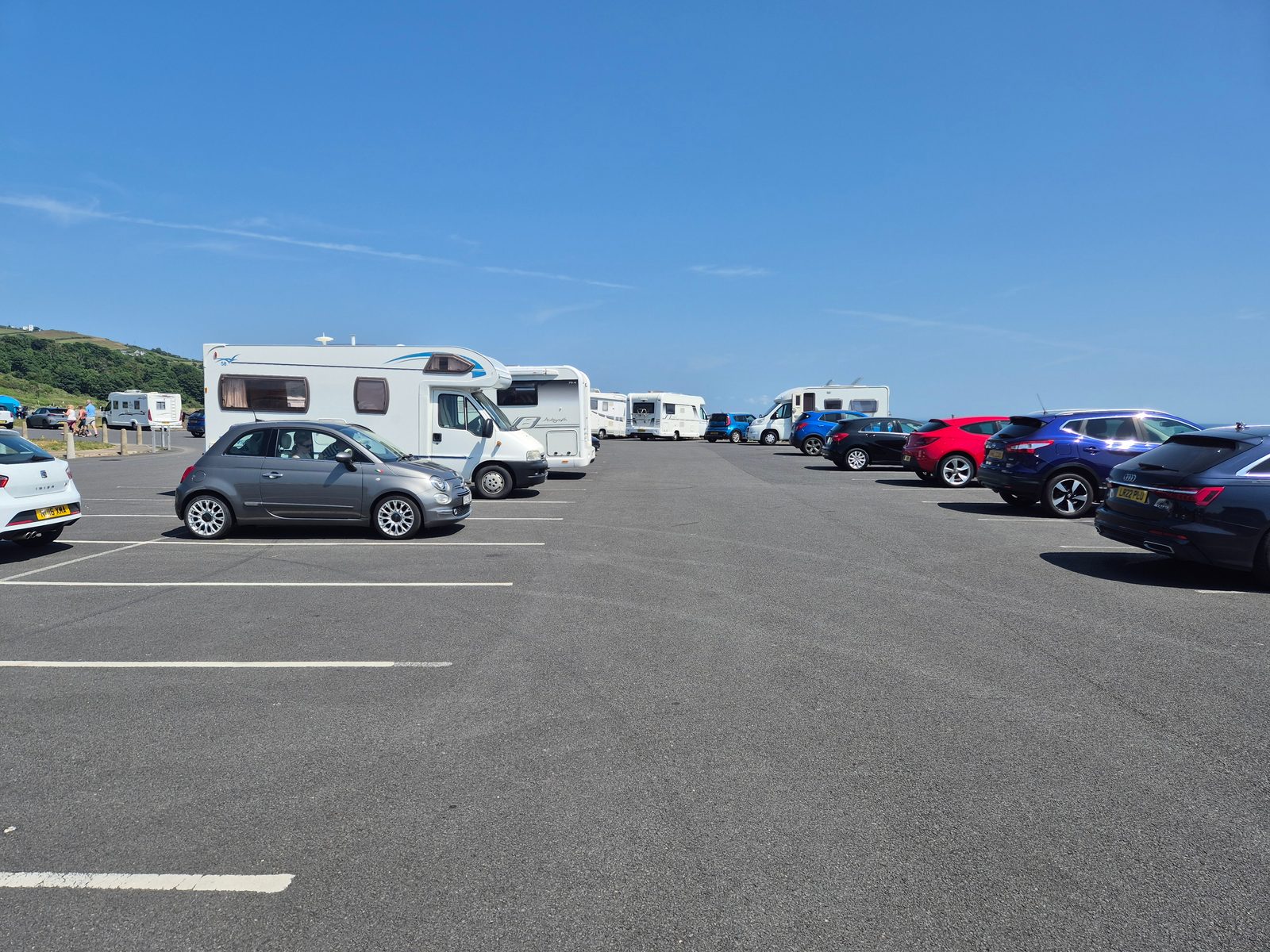 A tarmac car park with several coachbuilt motorhomes and cars including a grey Fiat 500 and a blue Nissan, with green hills and sea in the background under a blue sky.