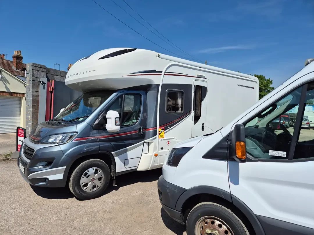 A grey and white Auto-Trail coachbuilt motorhome on a Fiat base parked next to a white panel van outside the garage under a blue sky.