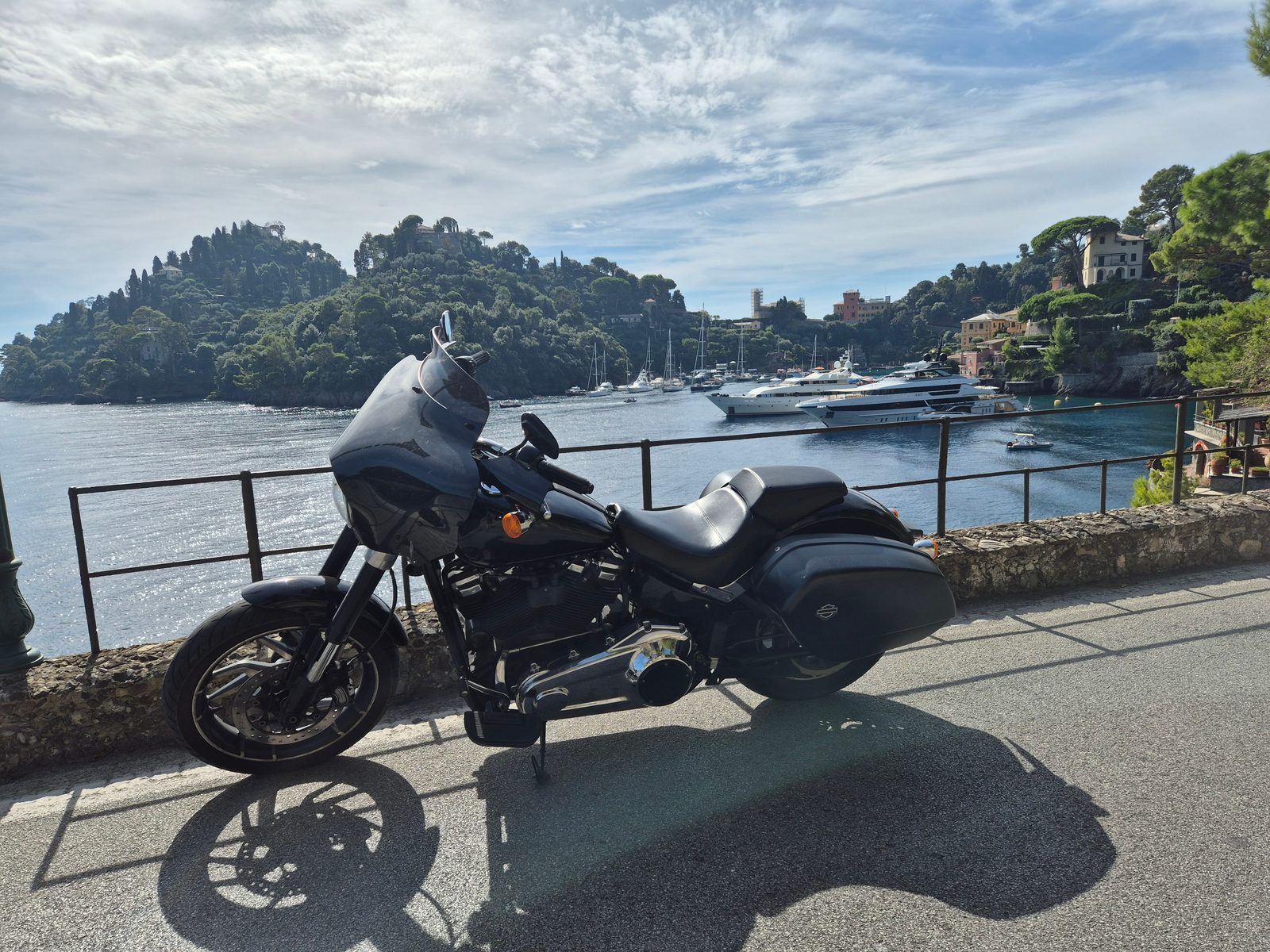 A black Harley-Davidson motorcycle parked by a railing overlooking a harbour with boats and yachts and hills behind.