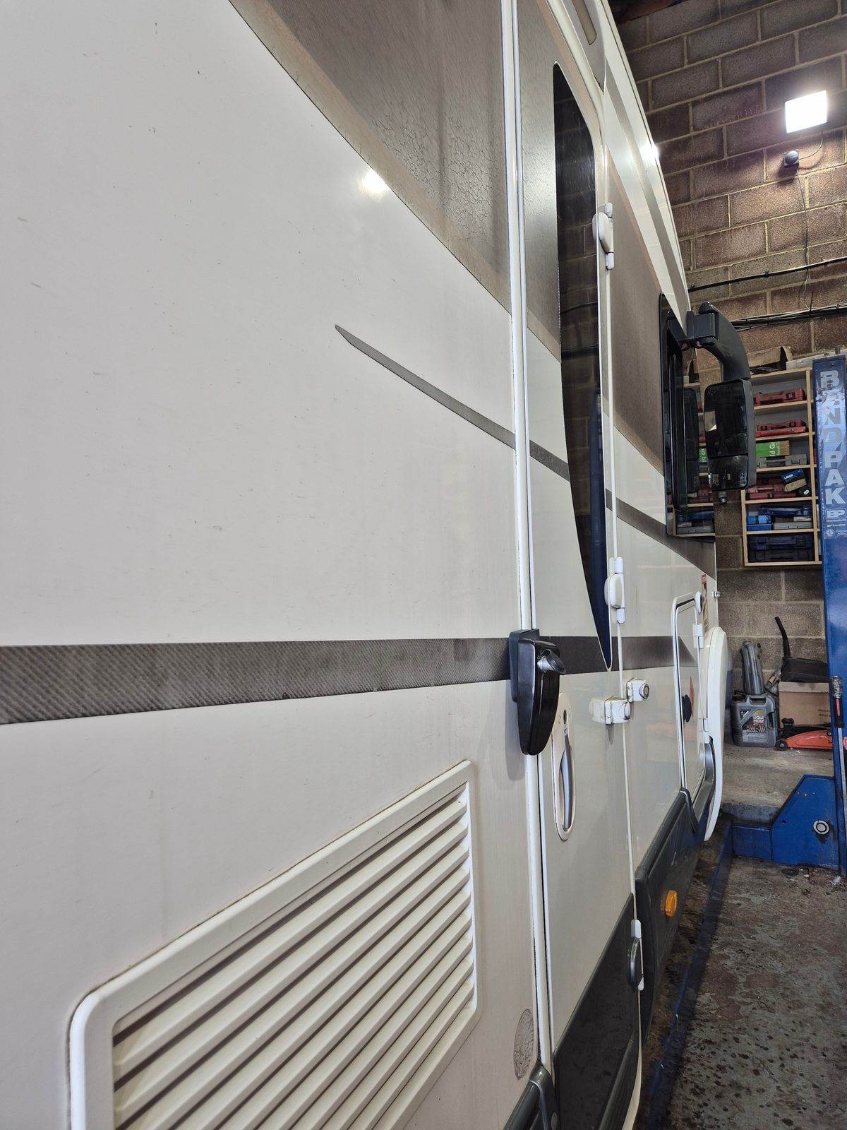 Side view of a white coachbuilt motorhome with a grey graphic stripe, external vent, door and wing mirror, parked next to a blue BendPak lift and shelving in a garage.