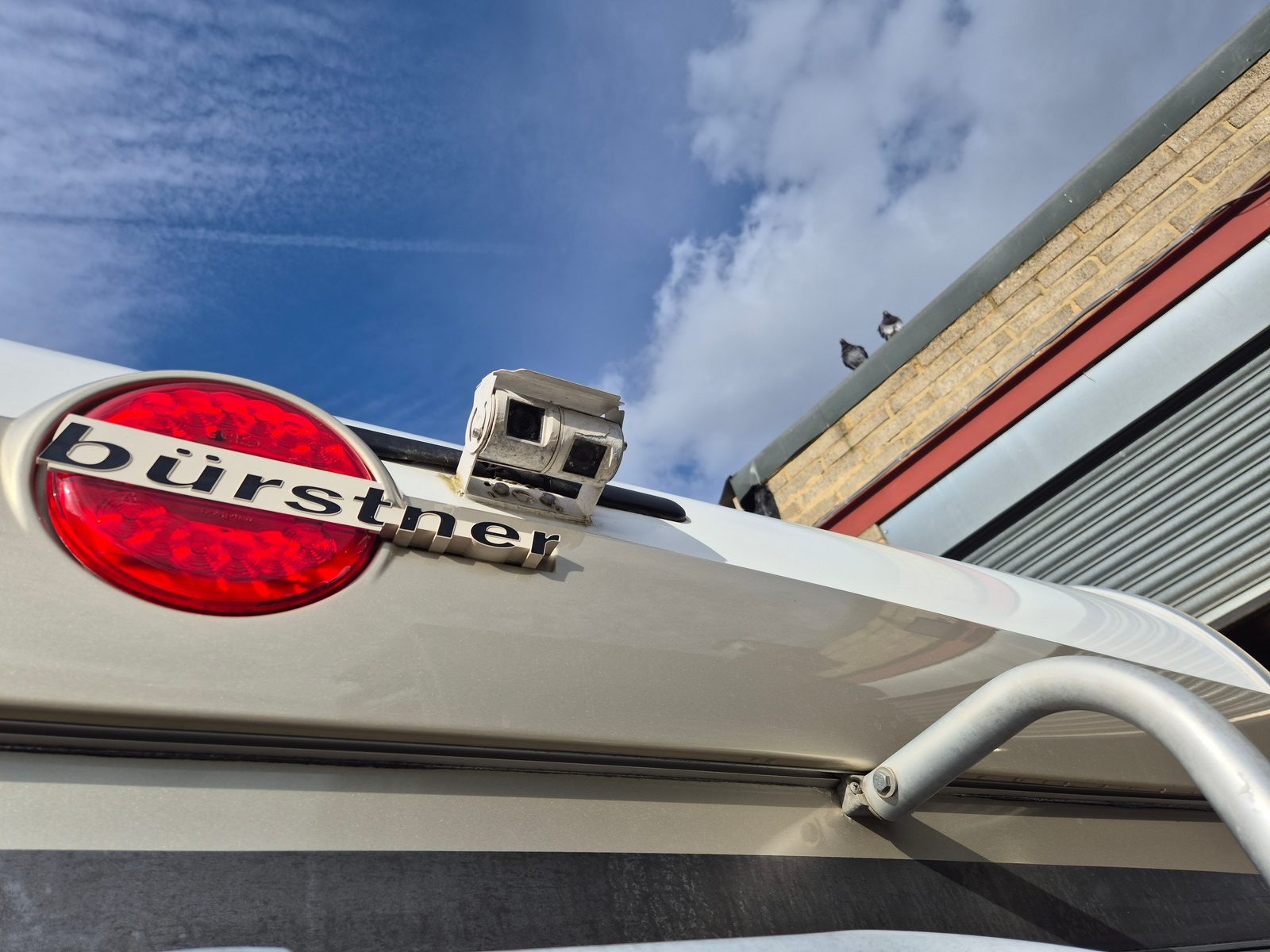 Close-up of a silver Bürstner motorhome's rear top corner, showing a round red tail light, badge and a twin-lens reversing camera against a blue sky.