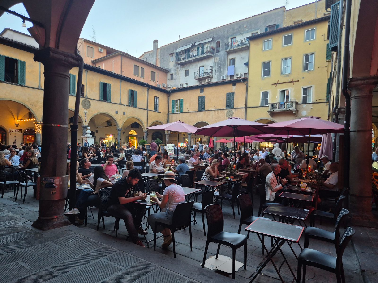 A busy evening in an Italian square with people sitting at outdoor tables under pink umbrellas, surrounded by old buildings with arched walkways.