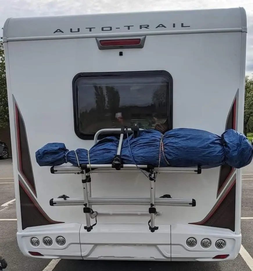 The back of a white Auto-Trail coachbuilt motorhome with a rear window and a cycle rack holding bikes wrapped in a blue cover, parked in a car park.