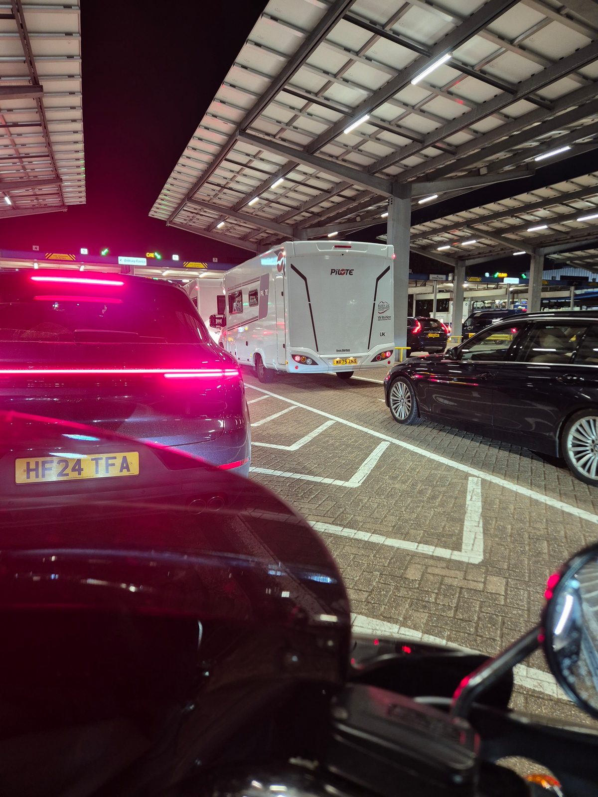 A white Pilote coachbuilt motorhome waiting in a queue of cars under a covered ferry terminal at night, with a dark SUV and estate car nearby.