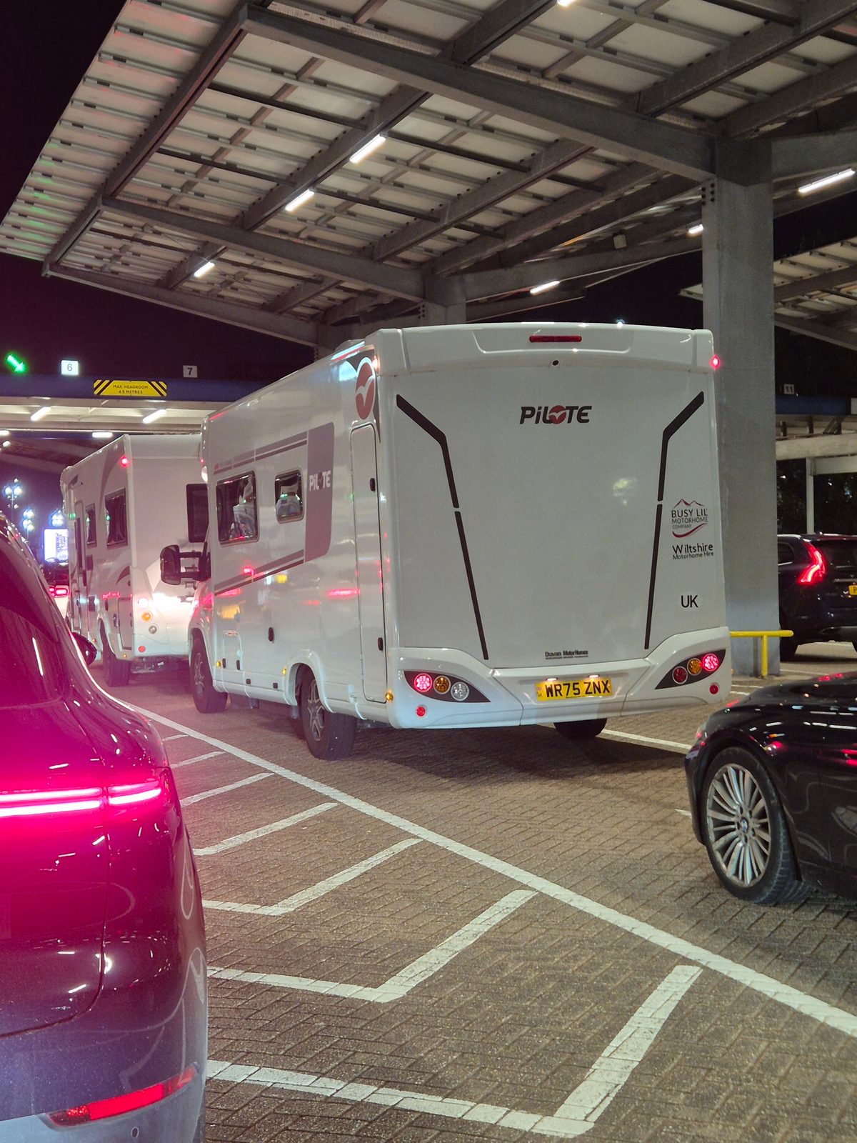 Rear view of a white Pilote coachbuilt motorhome, registration WR75 ZNX, queued between cars in a covered multi-storey car park at night.