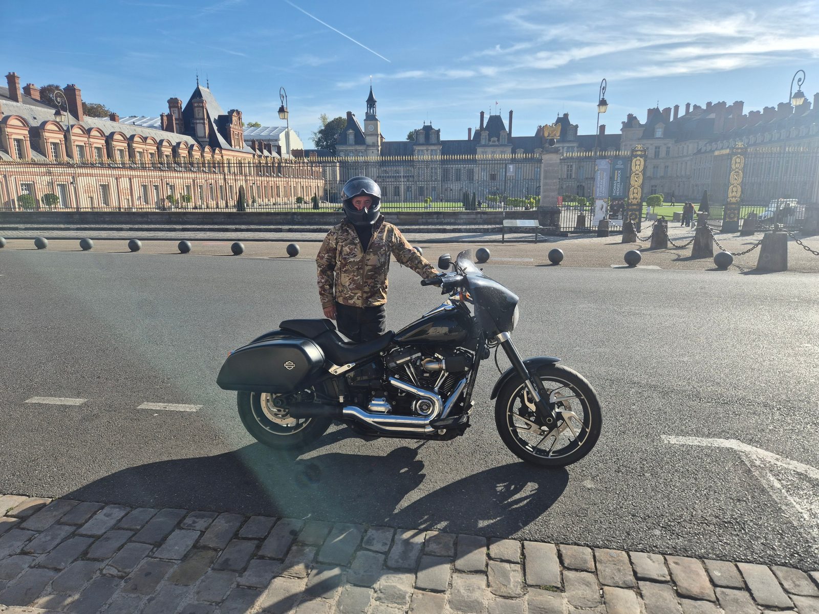 A man in a camouflage jacket and helmet standing beside a black Harley-Davidson outside the Palace of Fontainebleau in France.