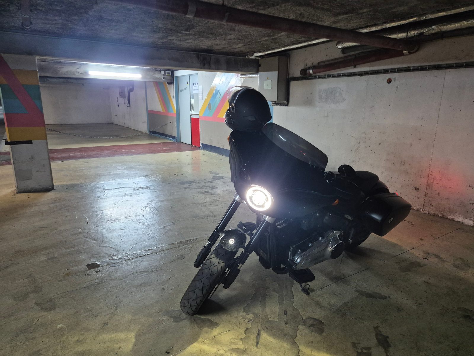 A black Harley-Davidson with a fairing and panniers parked in an underground car park with its headlight on.