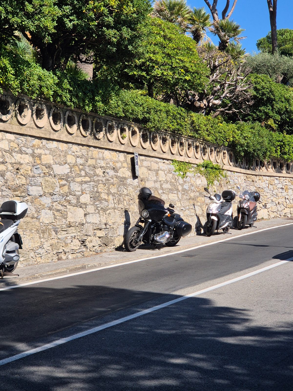 A black motorcycle and two scooters parked along a sunny street beside an ornate stone wall with trees above.