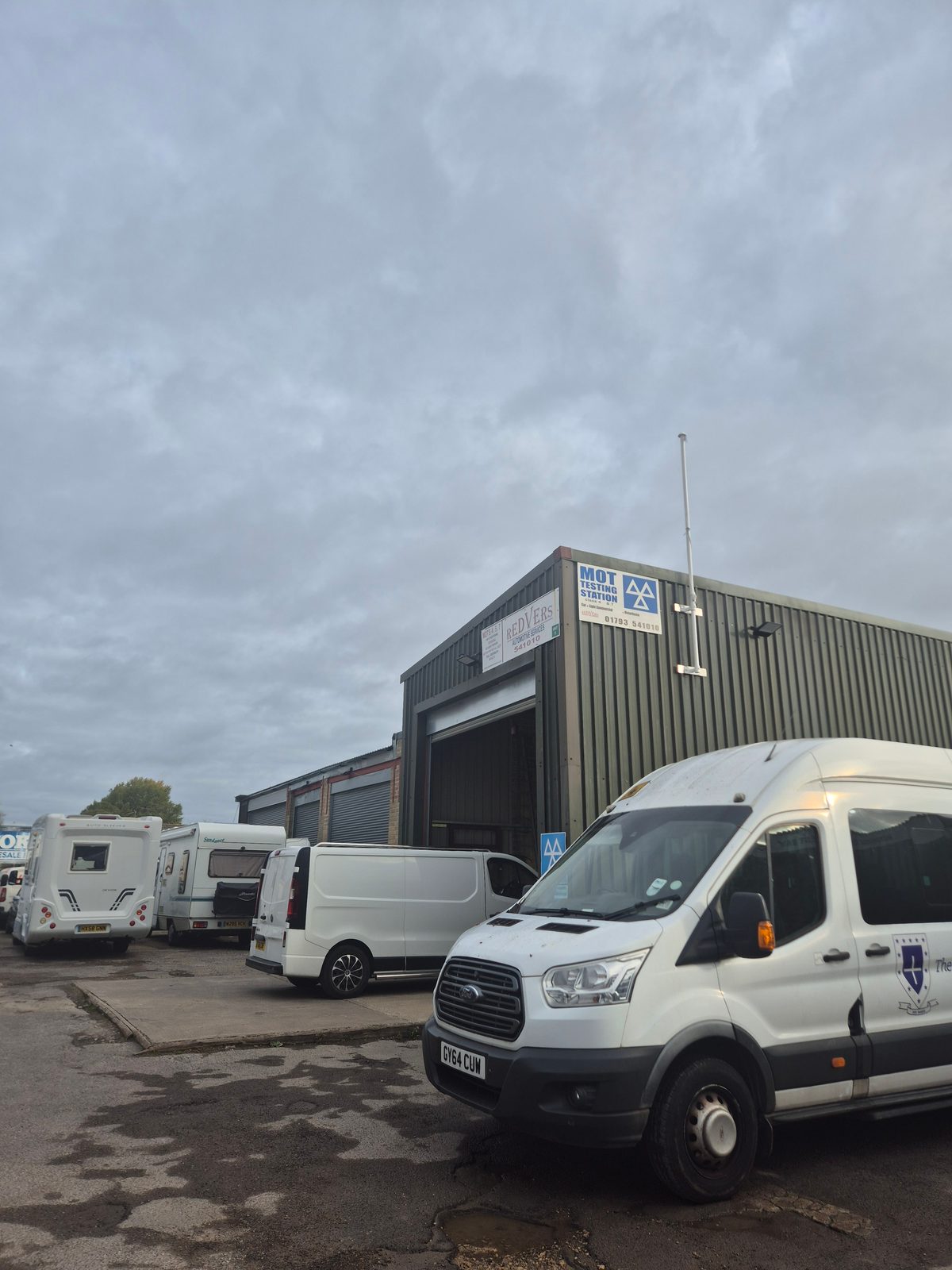 Several vehicles including a white minibus, a van and motorhomes parked outside the Redvers MOT testing station on a grey day.