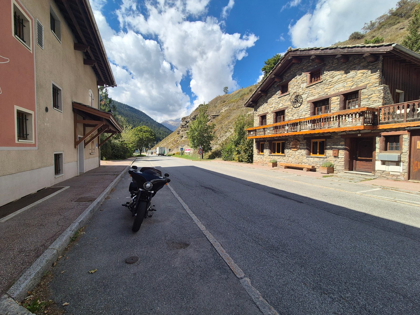 A black motorcycle parked on a quiet village street in the mountains with a stone chalet and green hillsides behind.