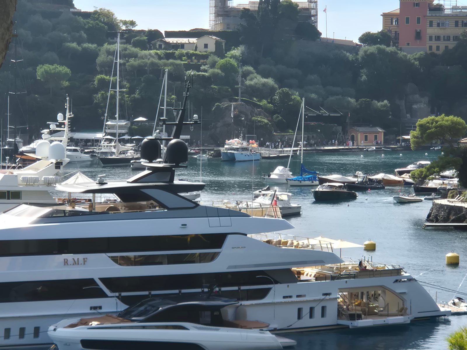 Large white superyachts and smaller boats moored in a harbour below a green hillside dotted with villas.