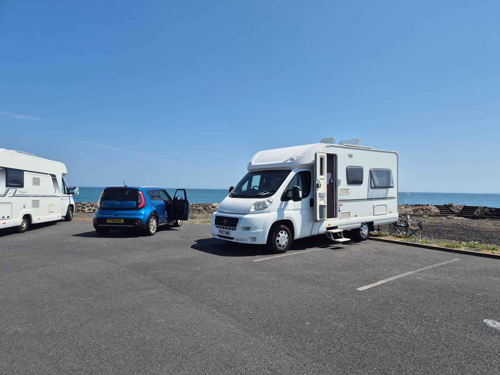 A white Fiat coachbuilt motorhome with its door open, a blue Kia Soul and a second white motorhome parked in a seafront car park under a clear blue sky.