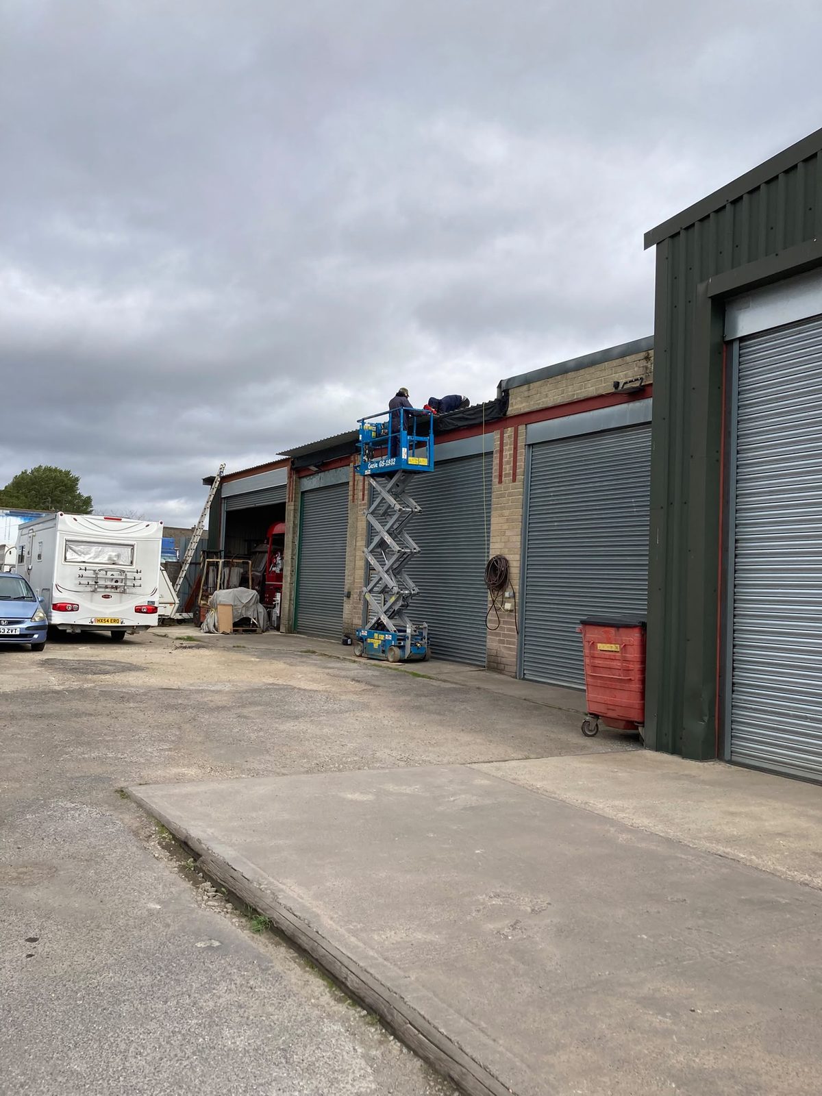 Two workers on a blue scissor lift working on the roof edge of an industrial garage unit with roller shutter doors and a motorhome parked nearby.