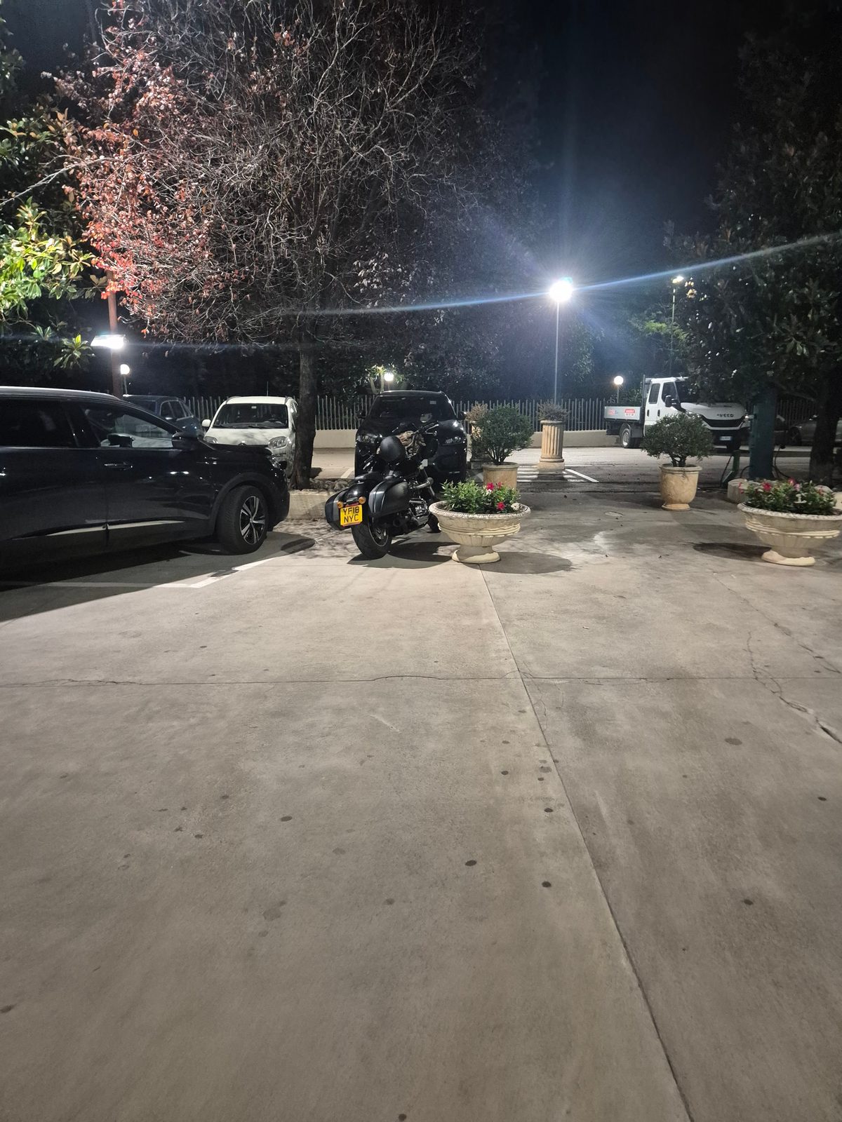A black motorcycle parked at night in a car park next to cars and stone planters, under a street light.