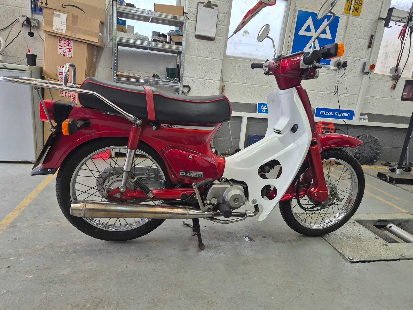 A red and white Honda Cub 90 moped on its stand inside a garage, with an MOT testing sign and shelving in the background.