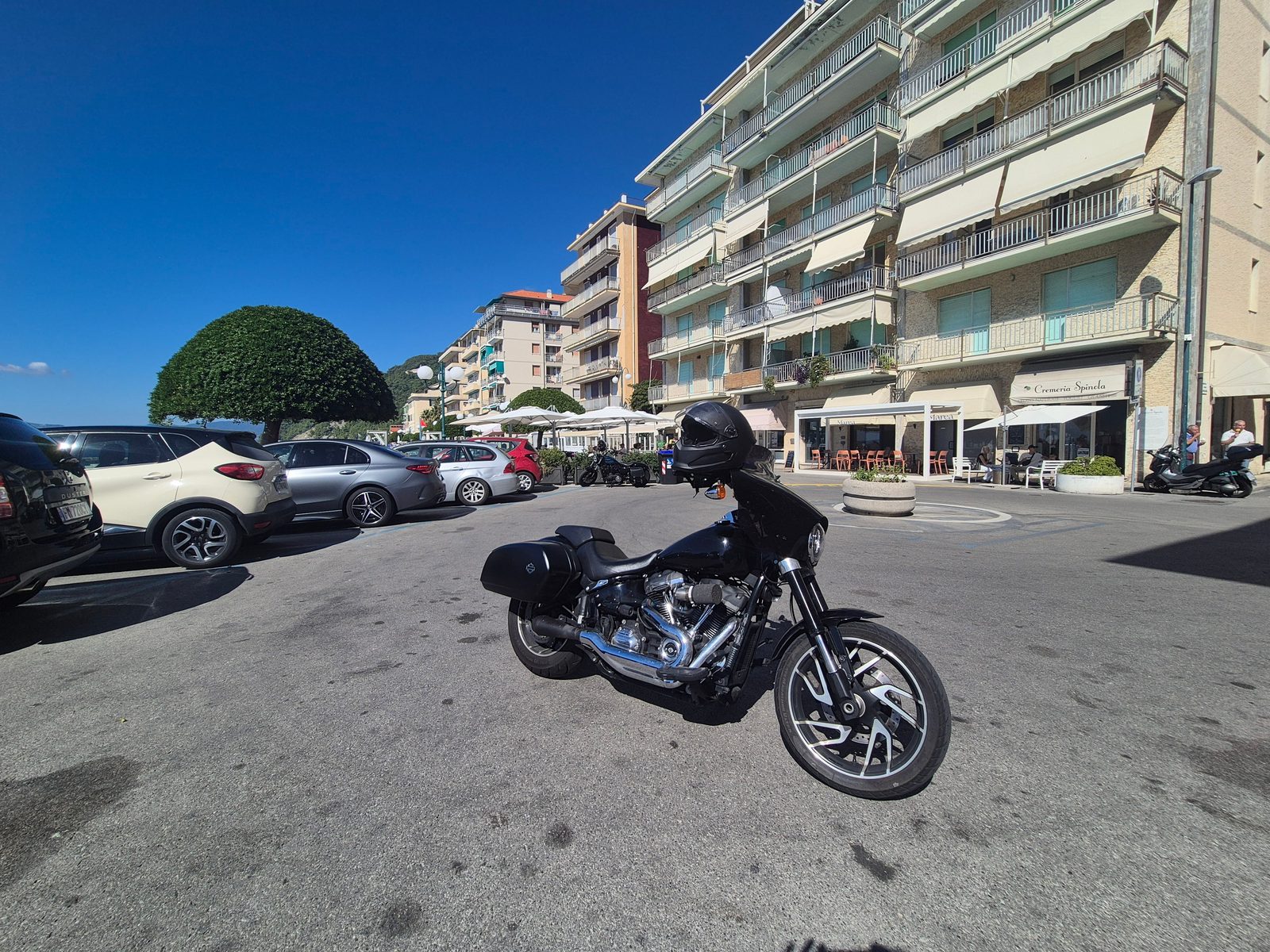 A black Harley-Davidson motorcycle parked in a sunny seaside town square with cafes and apartment buildings behind.