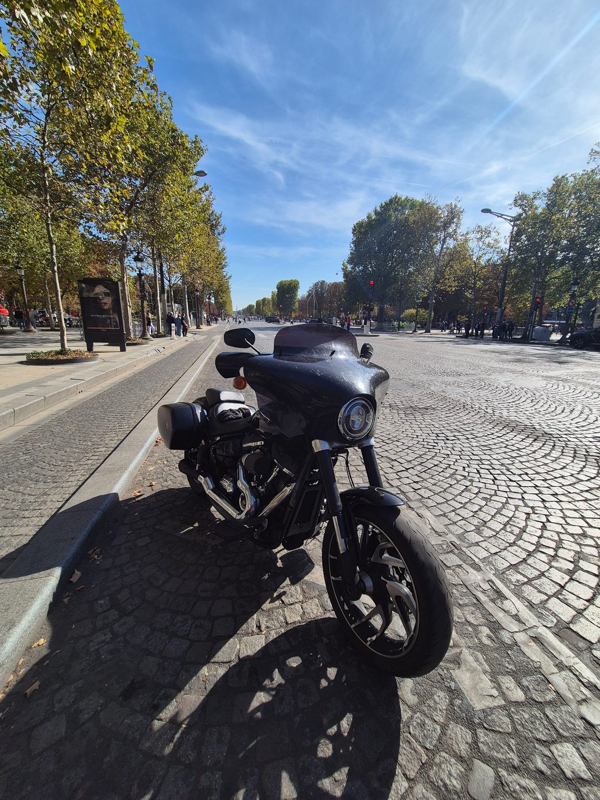 A black Harley-Davidson with a fairing parked on cobbles along a tree-lined Paris avenue.