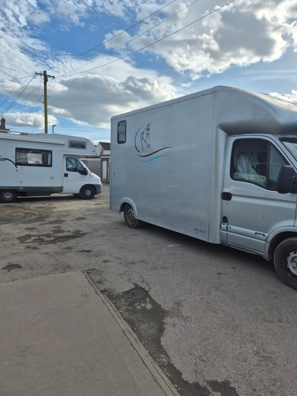 A silver horsebox van with a horse graphic parked beside a white coachbuilt motorhome on a tarmac yard under a cloudy sky.