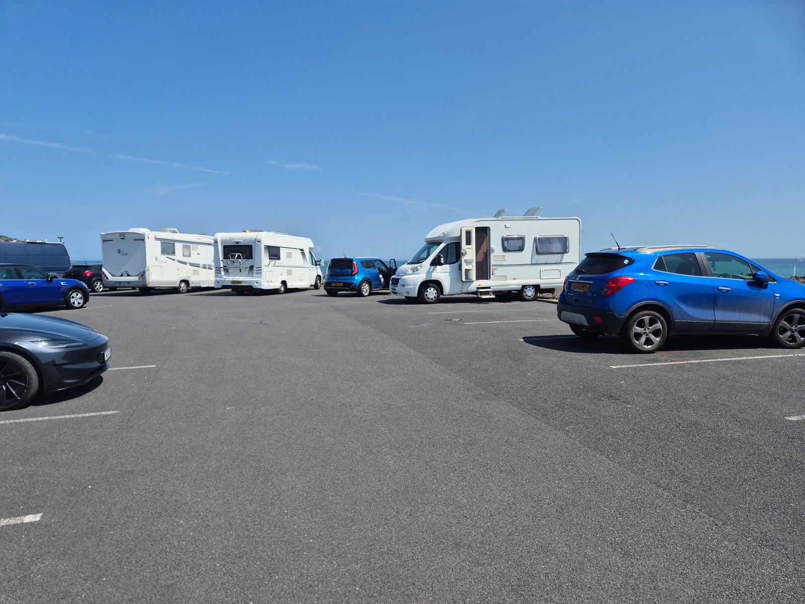 A tarmac car park beside the sea with several white motorhomes, a blue SUV, a grey saloon and other cars parked under a clear blue sky.