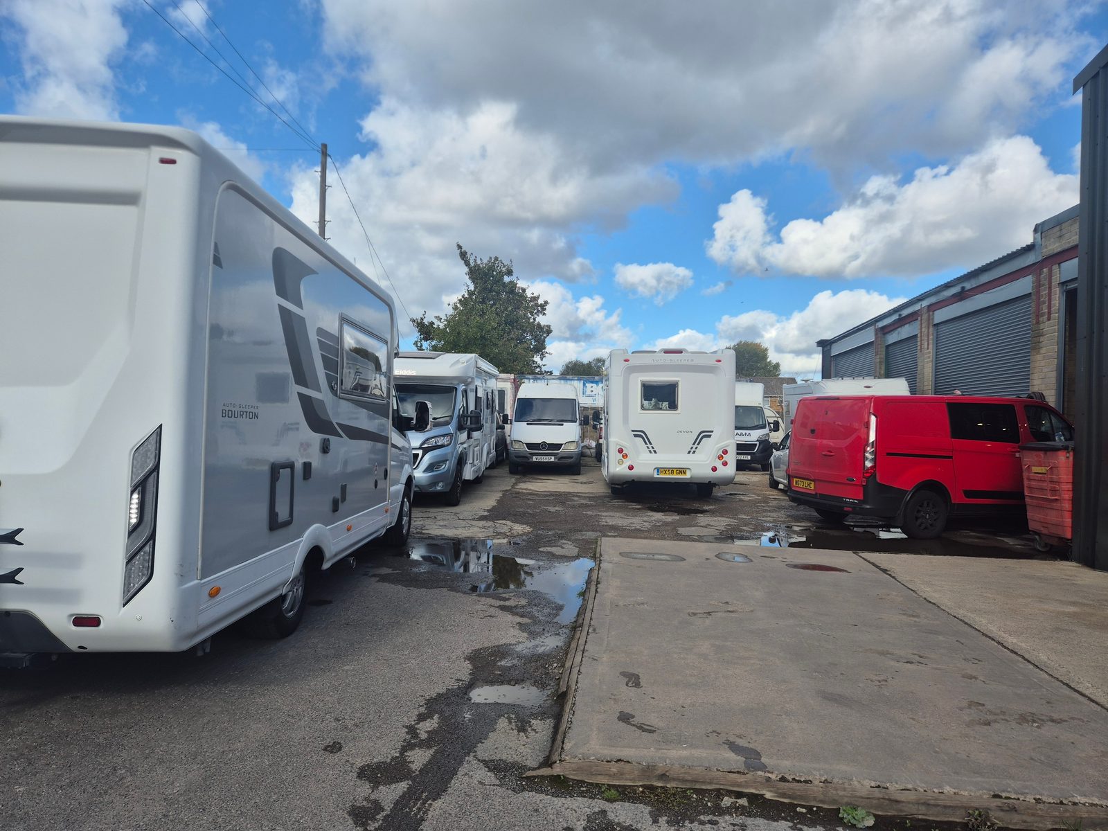 Several motorhomes and vans parked in a busy garage yard under a blue sky with clouds.