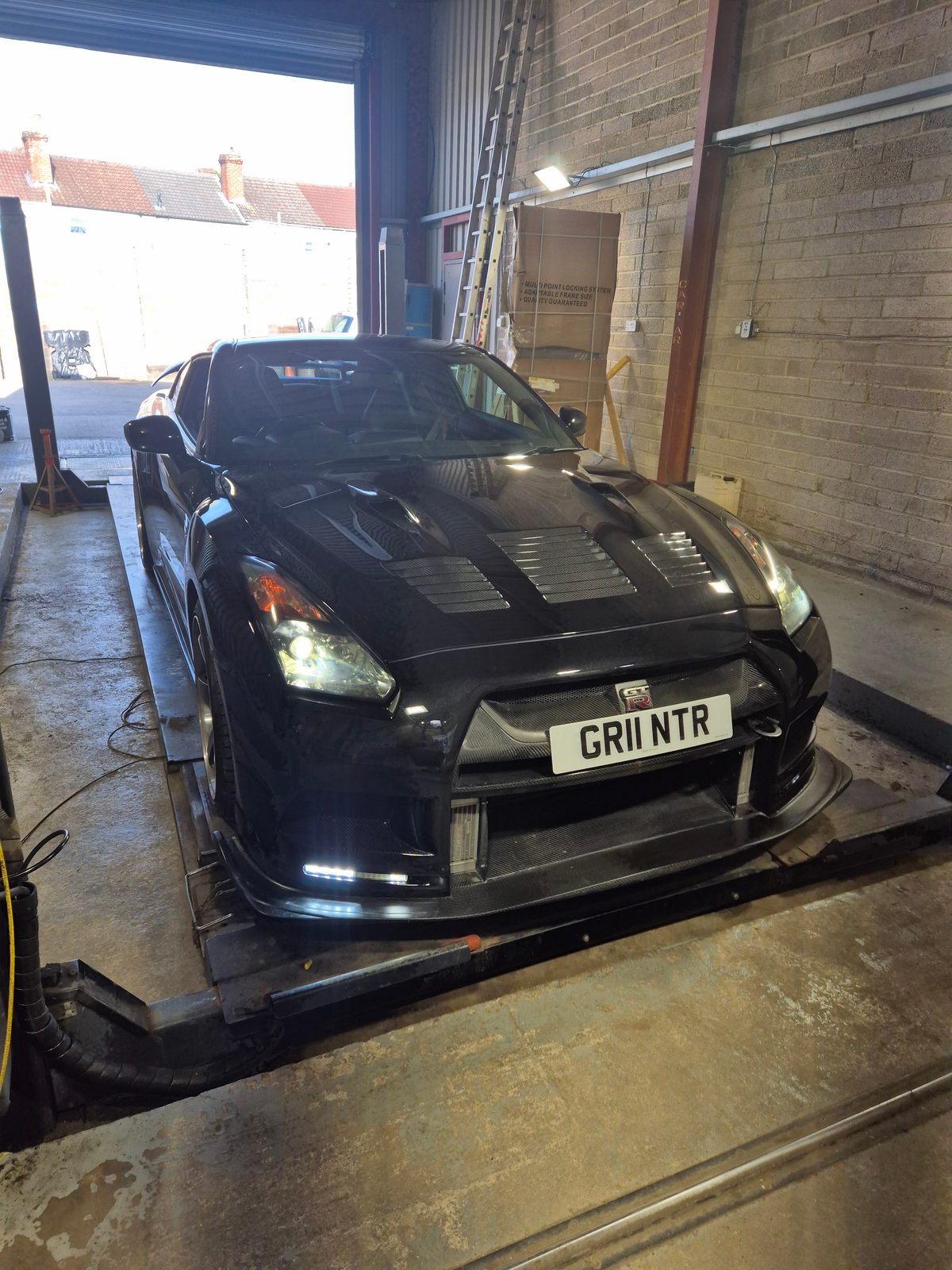 A black Nissan GT-R coupe with front number plate GR11 NTR parked on a garage ramp beside a brick wall and open shutter door.