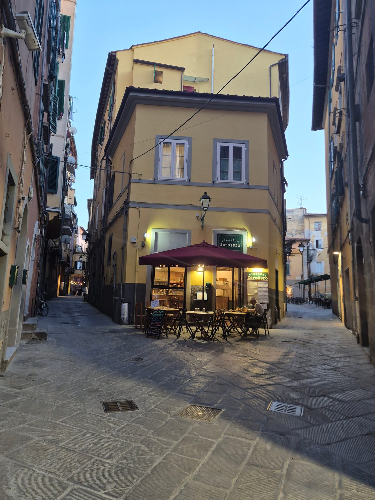 A small pizzeria with outdoor tables under a red umbrella on a narrow cobbled Italian street at dusk.