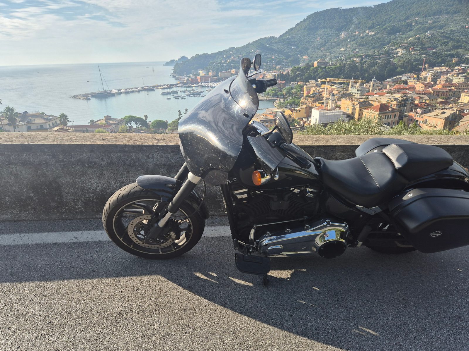 A black Harley-Davidson parked by a low wall overlooking an Italian coastal town and harbour full of boats.