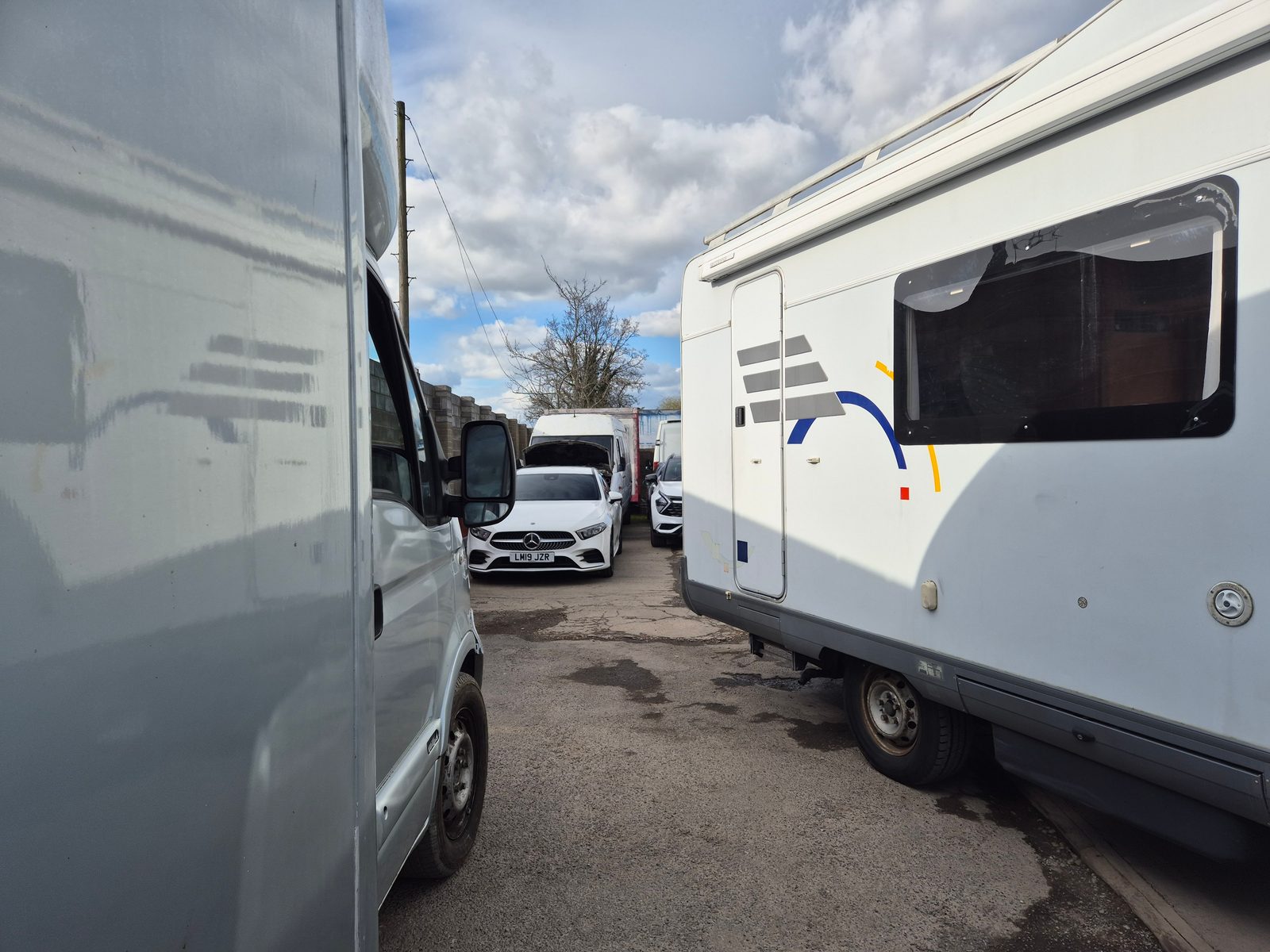 A white coachbuilt motorhome and a silver van parked close together in a garage yard, with a white Mercedes car and further vans visible between them under a cloudy sky.