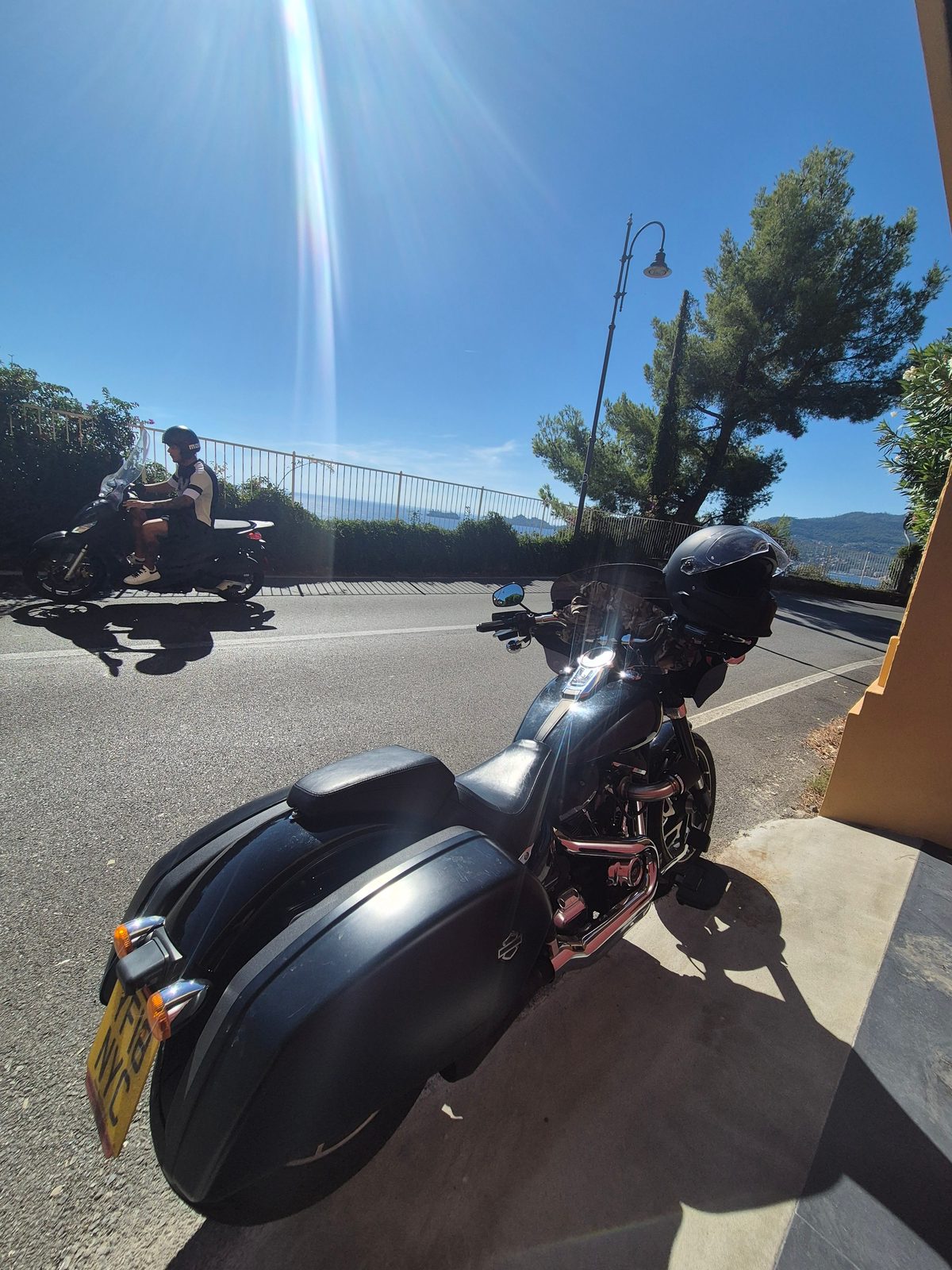 A black Harley-Davidson with panniers parked by a roadside overlooking the sea, with another rider passing on a scooter.