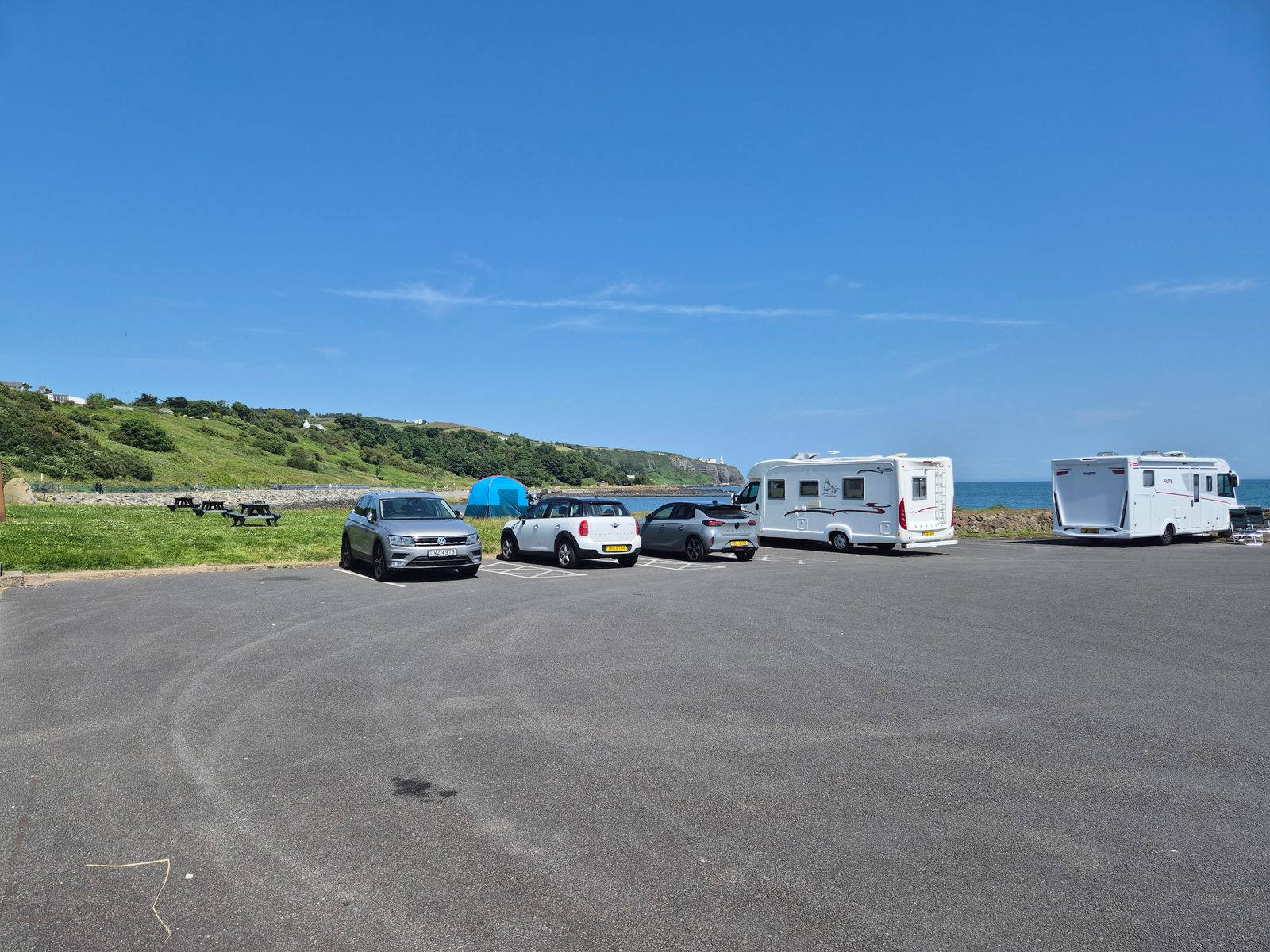 A seaside tarmac car park with two white motorhomes, a silver Volkswagen SUV, a Mini and a hatchback, plus picnic benches, a tent and grassy coastline under a clear blue sky.