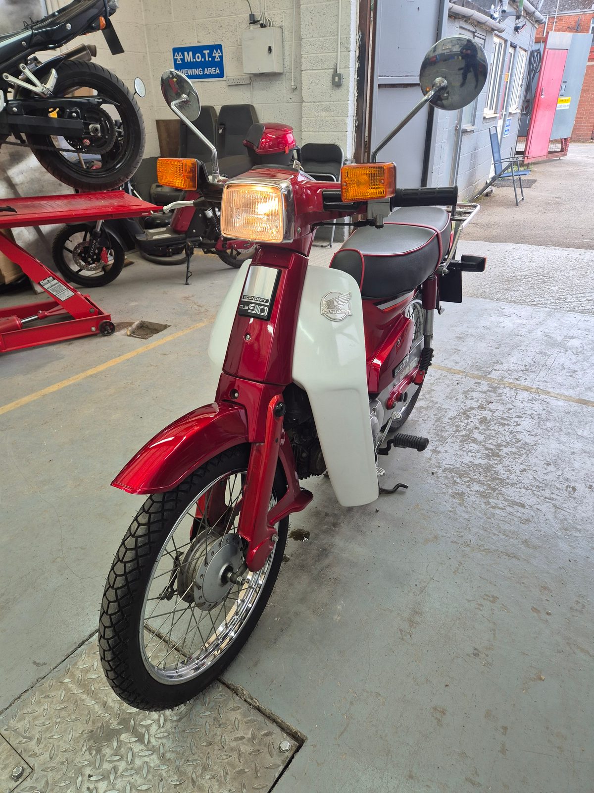 A red and white Honda Cub 90 step-through motorcycle parked indoors near an MOT viewing area, with another motorcycle on a ramp behind.