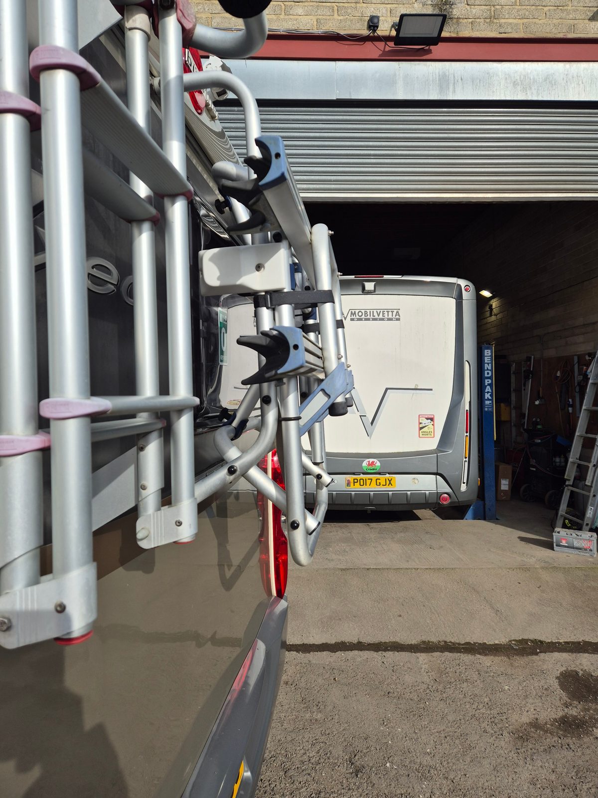 A white Mobilvetta coachbuilt motorhome with a Welsh flag sticker reversed into a garage workshop beside a blue BendPak two-post lift, viewed past a rear-mounted ladder and bike rack of another motorhome.