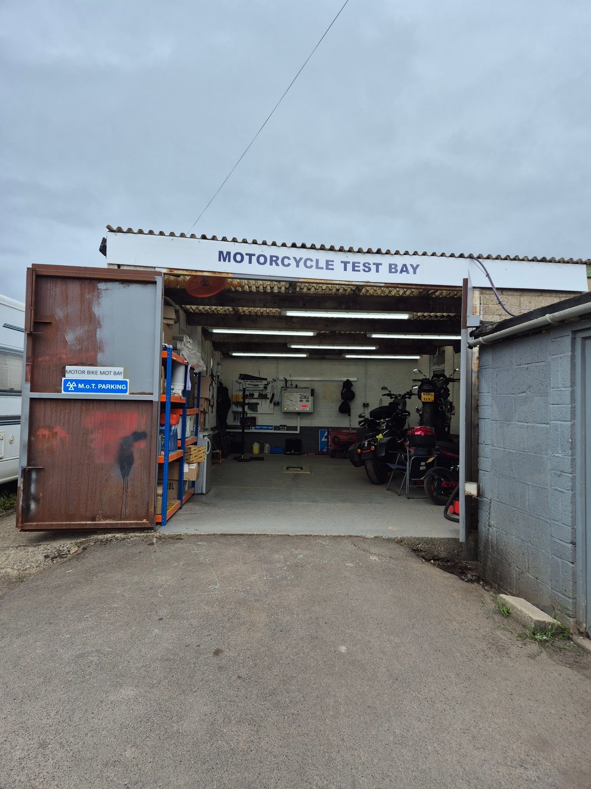 Open doors of a motorcycle MOT test bay with two motorcycles parked inside and tools on the wall.