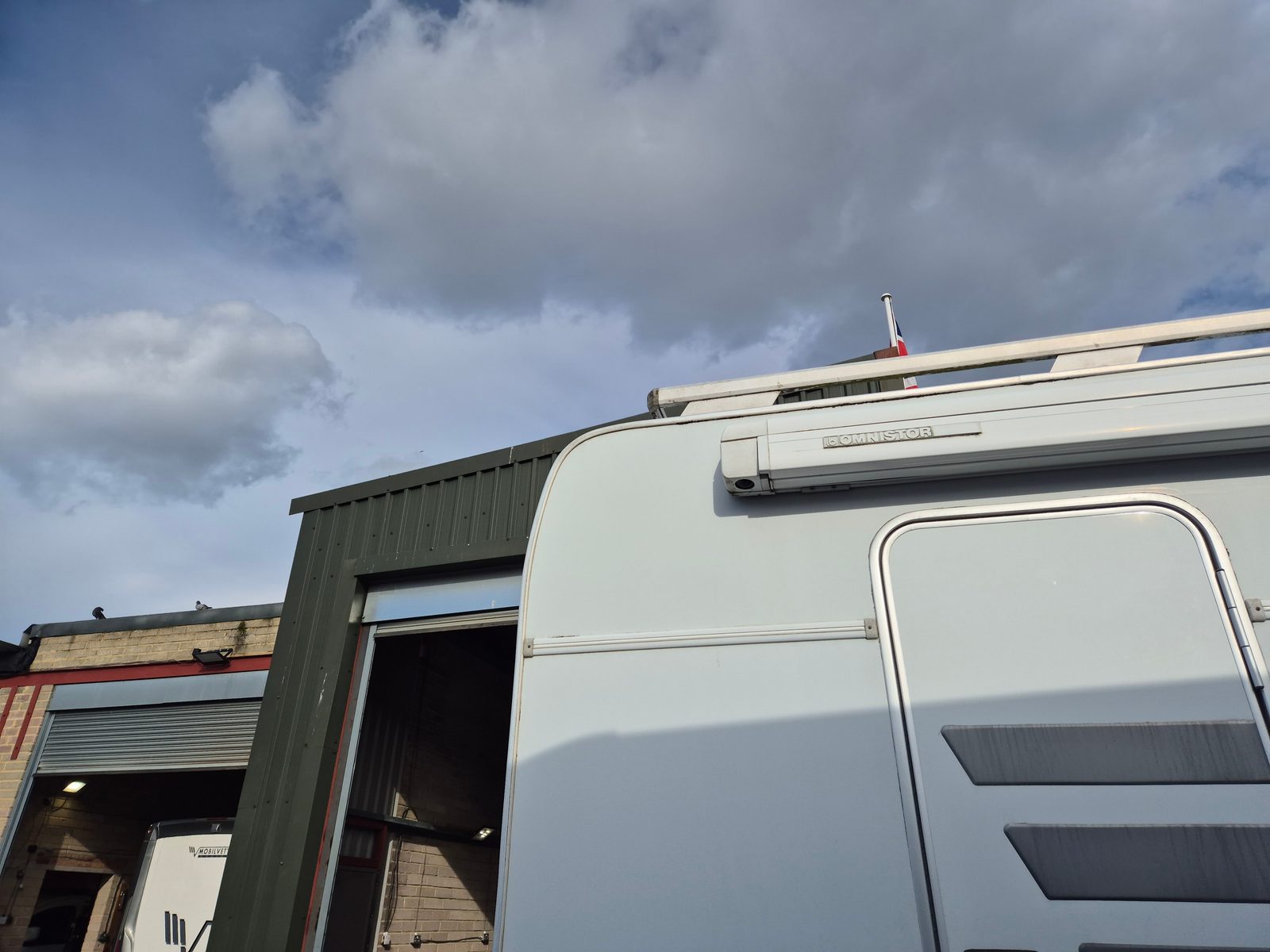 The upper side and rear of a white motorhome fitted with an Omnistor awning and roof rail, parked beside a workshop with an open roller shutter door under a cloudy sky.