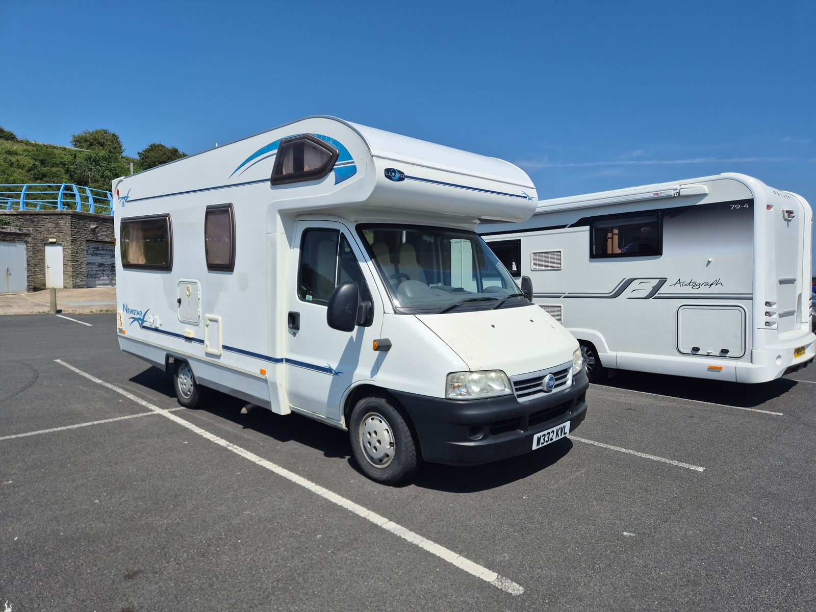 A white Lunar Newstar coachbuilt motorhome on a Fiat base beside a Bessacarr Autograph motorhome in a tarmac car park under a clear blue sky.