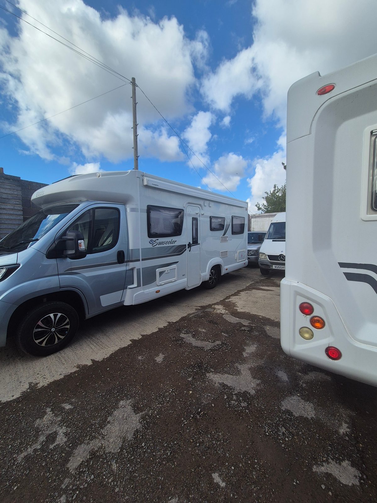 Two white coachbuilt motorhomes parked in a garage yard under a blue sky with scattered clouds.
