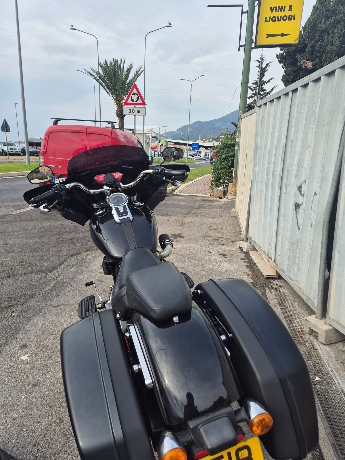 A black motorcycle with panniers parked by a road near a border crossing, with a red van, road signs and hills in the background.