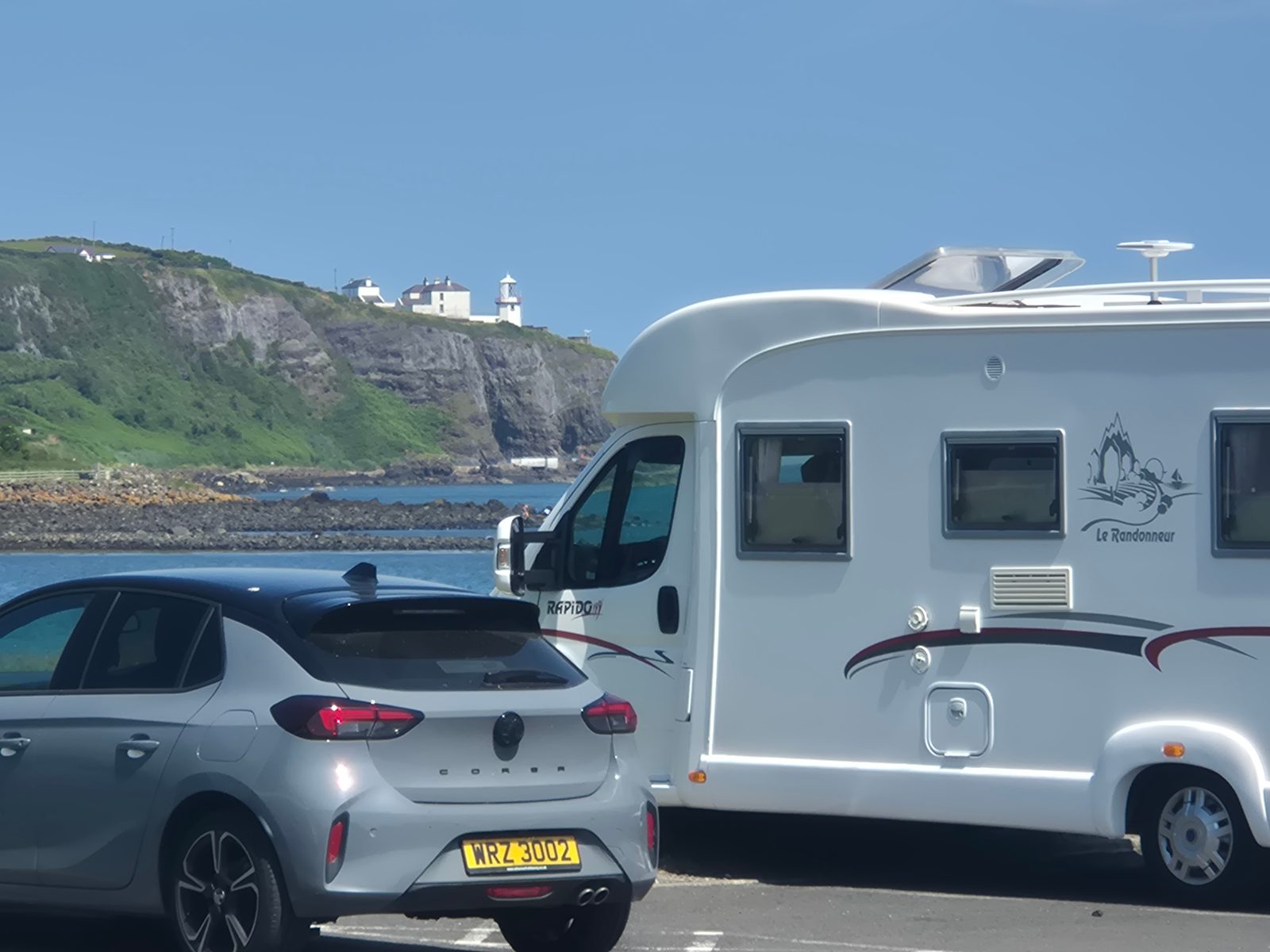 A silver Vauxhall Corsa and a white Rapido Le Randonneur motorhome parked by the sea, with a clifftop lighthouse in the background.