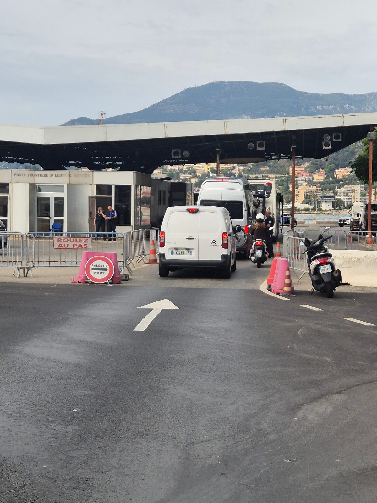 A police border checkpoint by the sea with vans, a scooter and traffic queuing under a large canopy.
