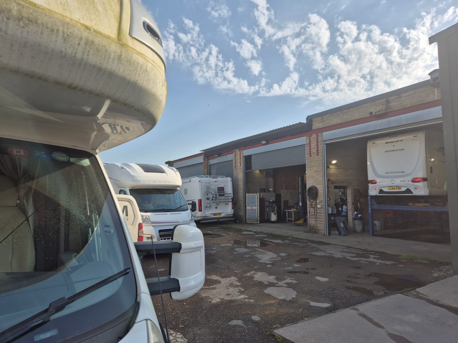 A row of coachbuilt motorhomes parked outside brick garage units, with a white Swift motorhome raised on a ramp inside an open workshop bay.