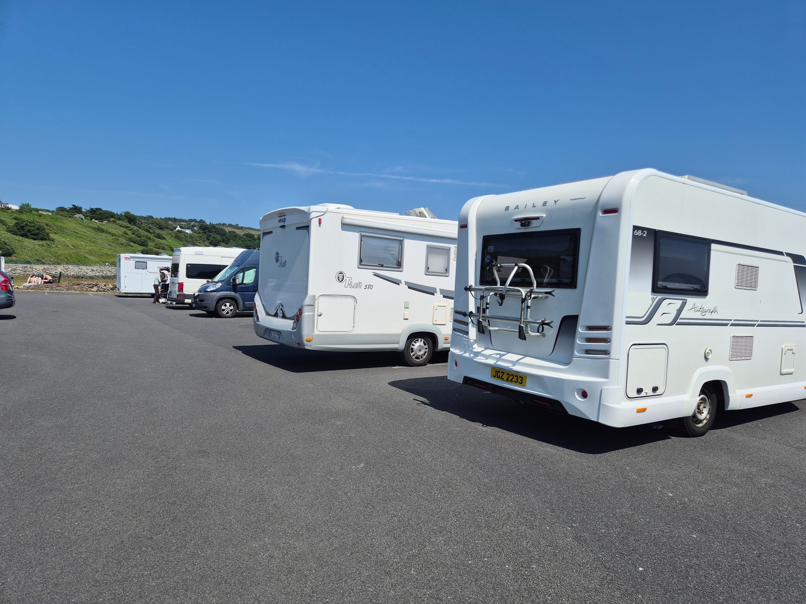 A white Bailey Autograph motorhome and an Elnagh T-Loft coachbuilt motorhome parked on tarmac, with a campervan and other vehicles nearby and green hills under a blue sky.