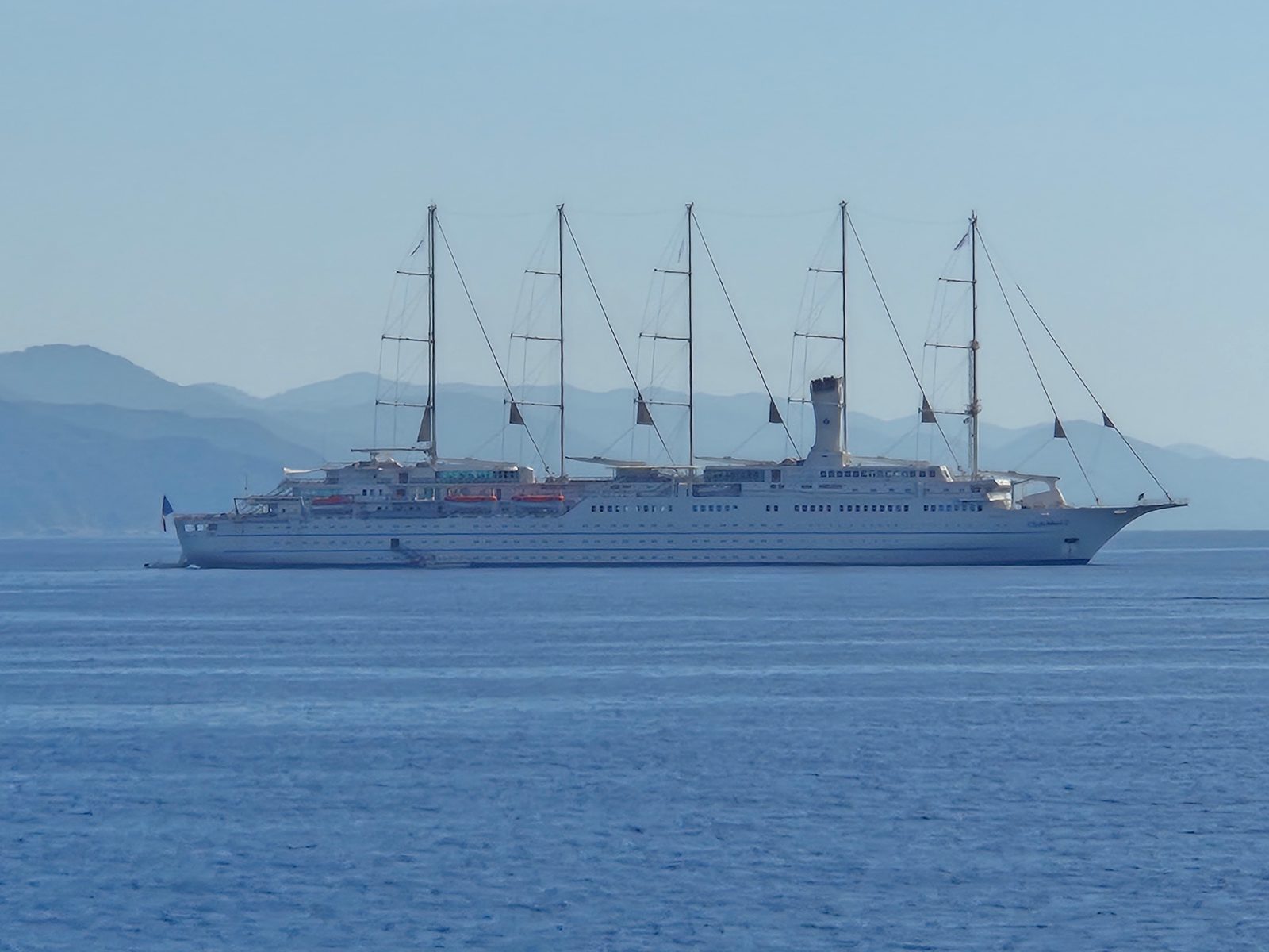 A large white five-masted cruise sailing ship anchored on calm water with hazy mountains behind.
