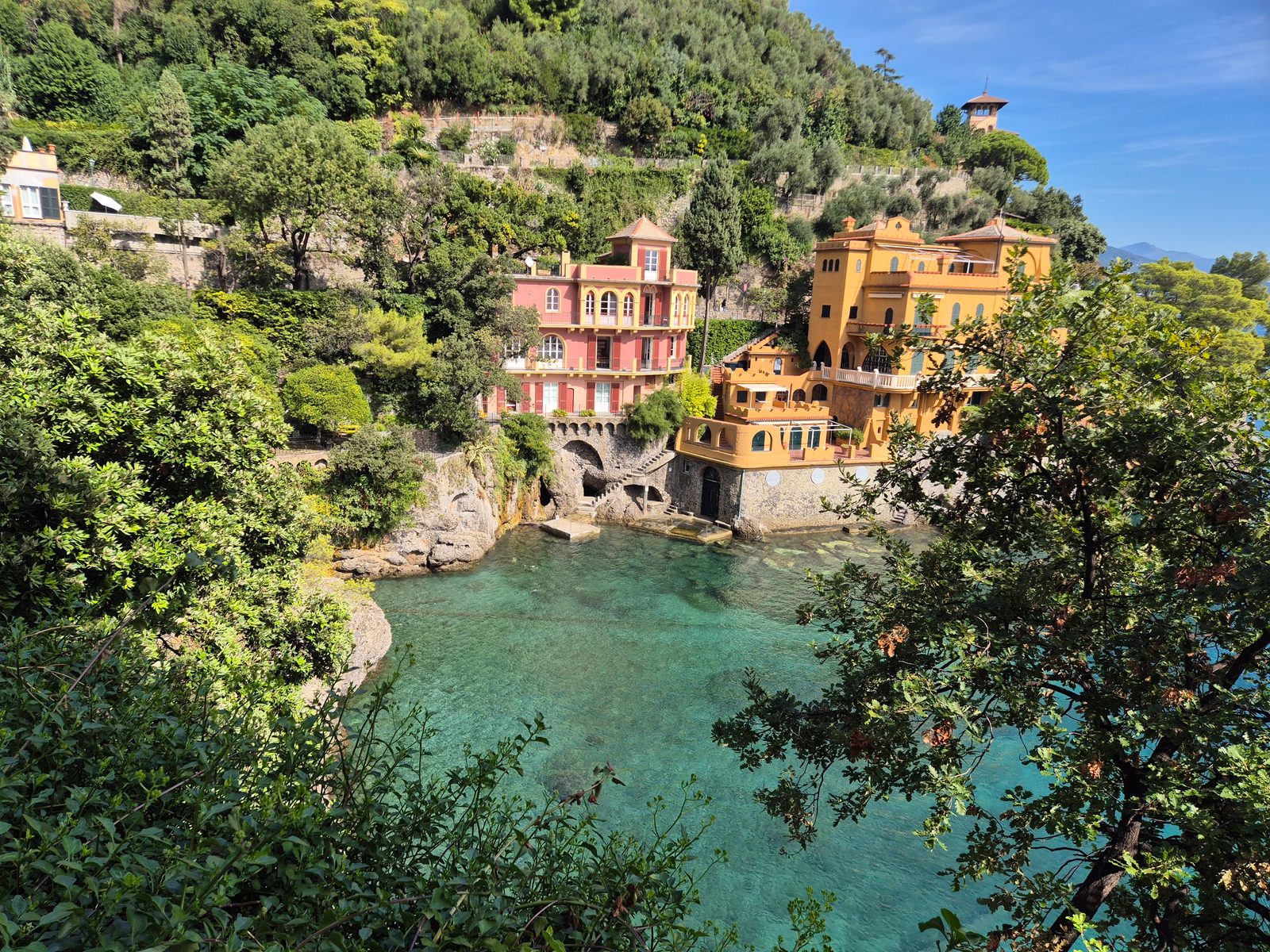 A sheltered cove with turquoise water and two grand villas built into a wooded hillside above the shore.