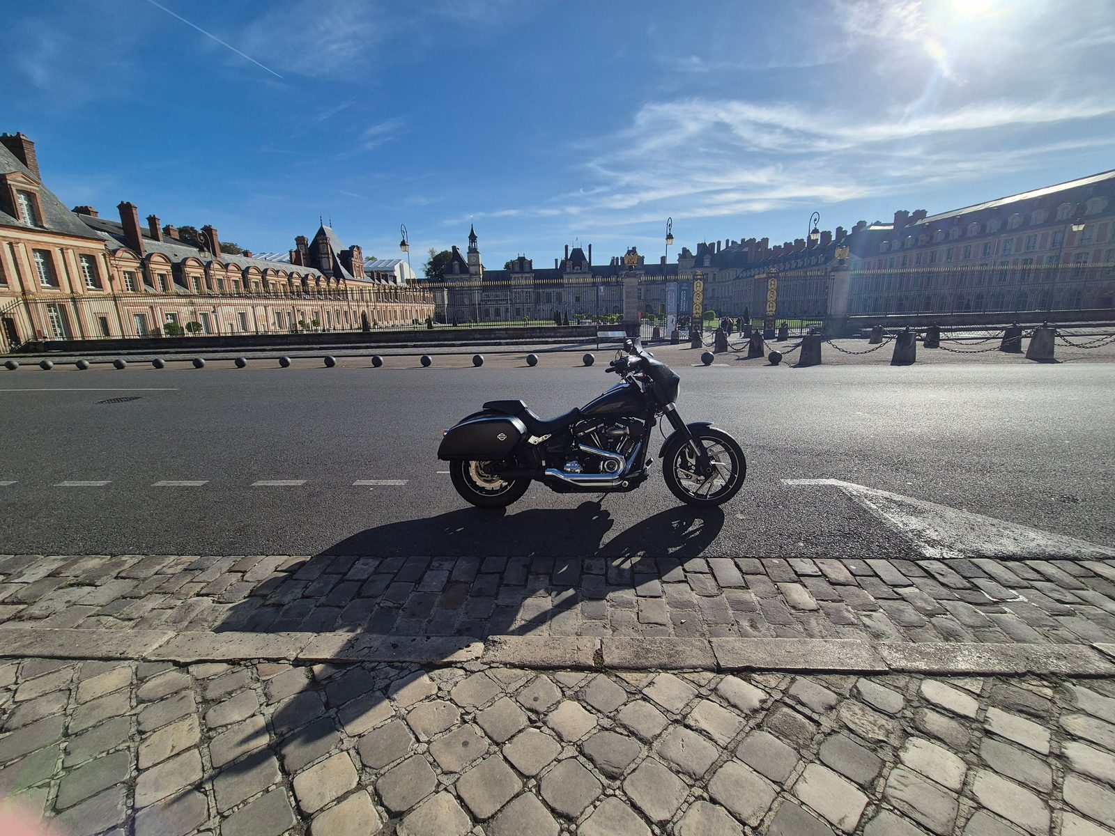 A black Harley-Davidson motorcycle parked on a cobbled road in front of the Palace of Fontainebleau under a blue sky.